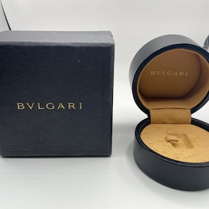 Puede incluir: Caja de joyería negra y estuche redondo con el nombre de la marca "BVLGARI" en letras doradas. El estuche redondo está abierto, revelando un interior beige. La caja es rectangular y cerrada.