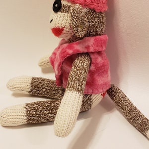May include: A brown and white knitted sock monkey wearing a pink crocheted hat with a white pom-pom and a pink fleece jacket.