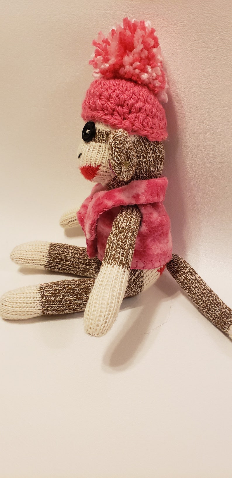 May include: A brown and white knitted sock monkey wearing a pink crocheted hat with a white pom-pom and a pink fleece jacket.