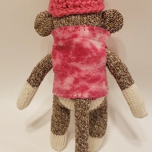 May include: A brown and white knitted sock monkey wearing a pink crocheted hat with a white pom-pom and a pink fleece scarf.