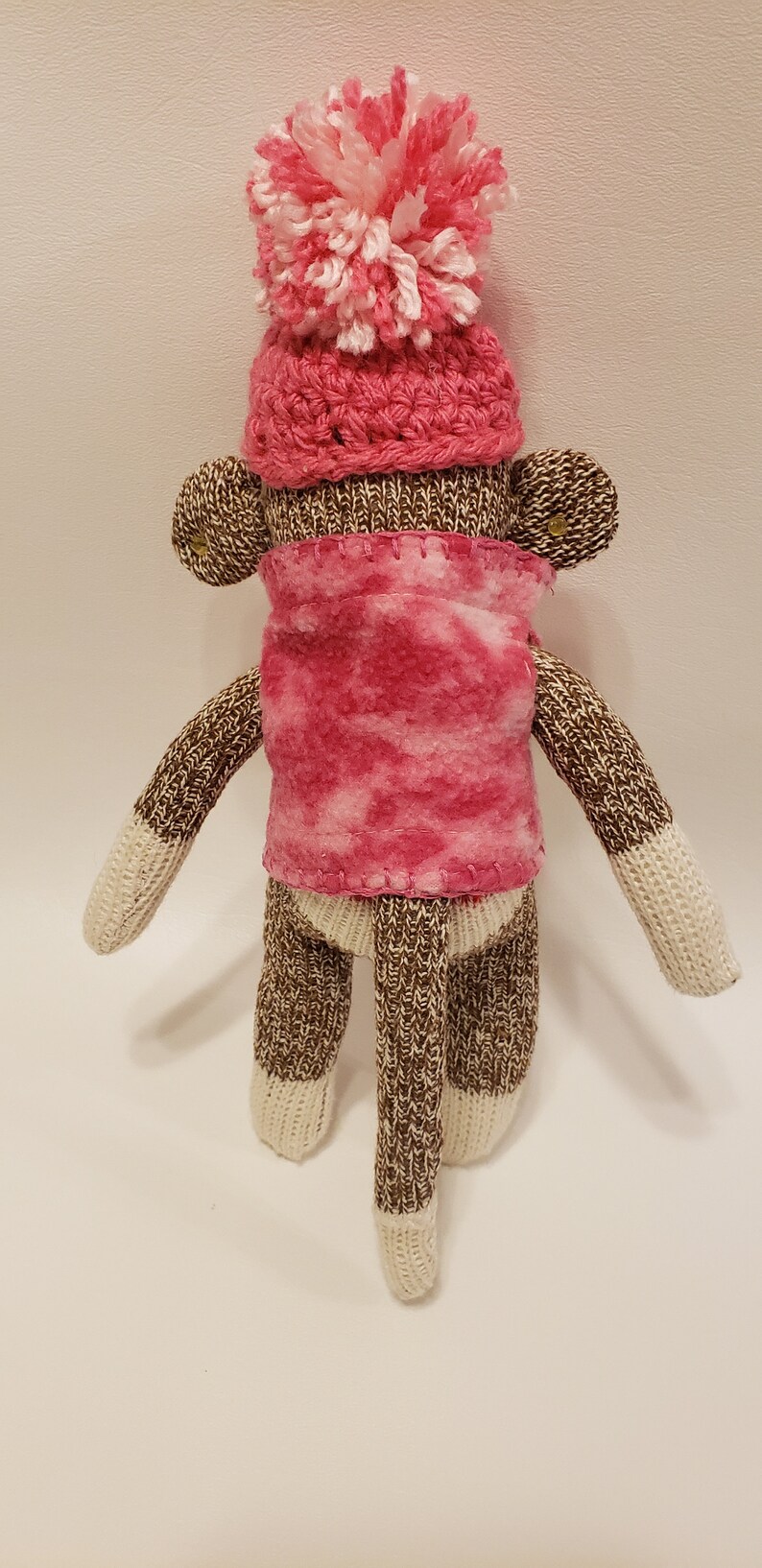 May include: A brown and white knitted sock monkey wearing a pink crocheted hat with a white pom-pom and a pink fleece scarf.