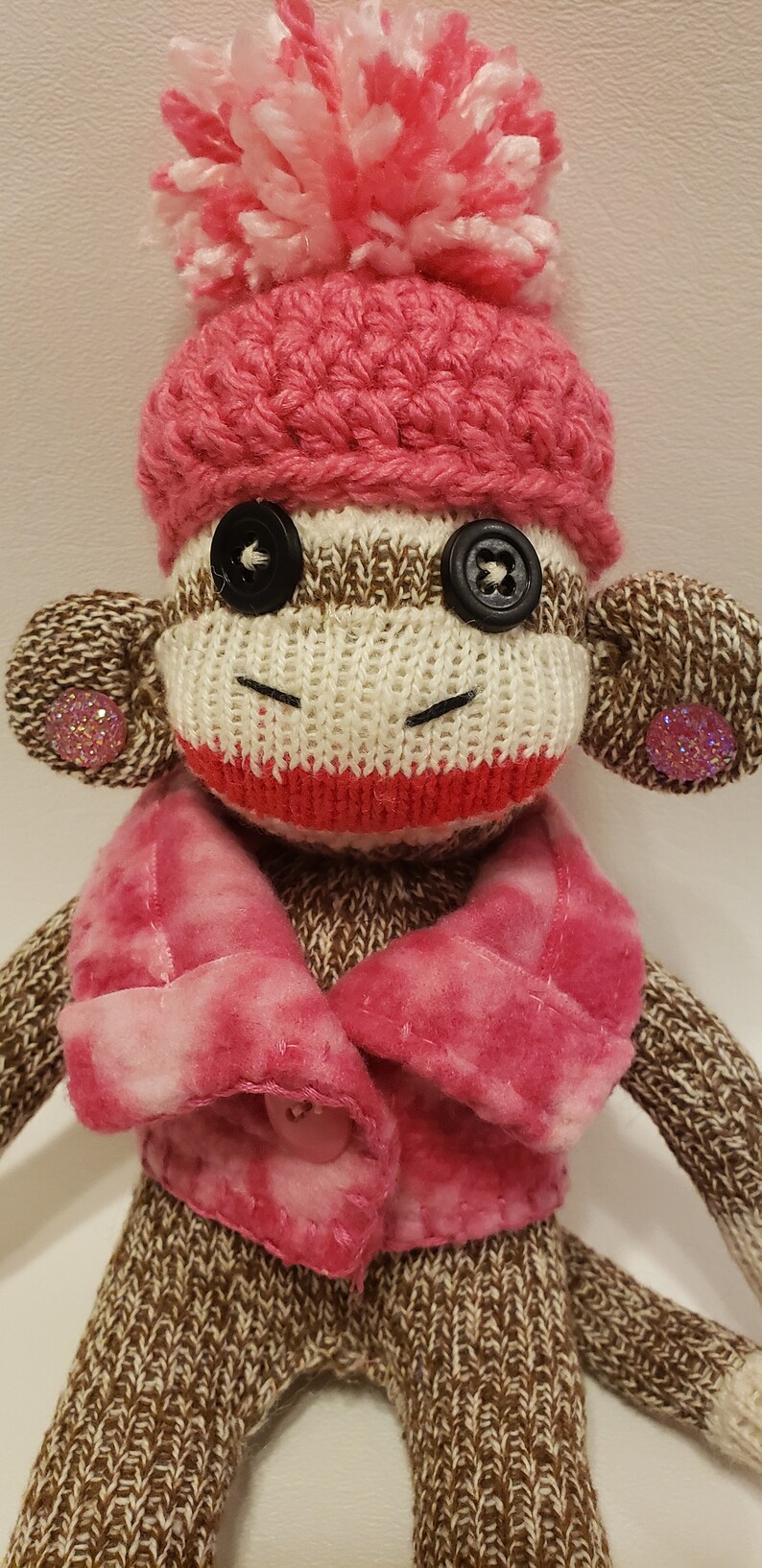 May include: A sock monkey wearing a pink crocheted hat with a white pom-pom, a pink fleece jacket, and brown knit pants. The monkey has black button eyes and pink glitter on its ears.