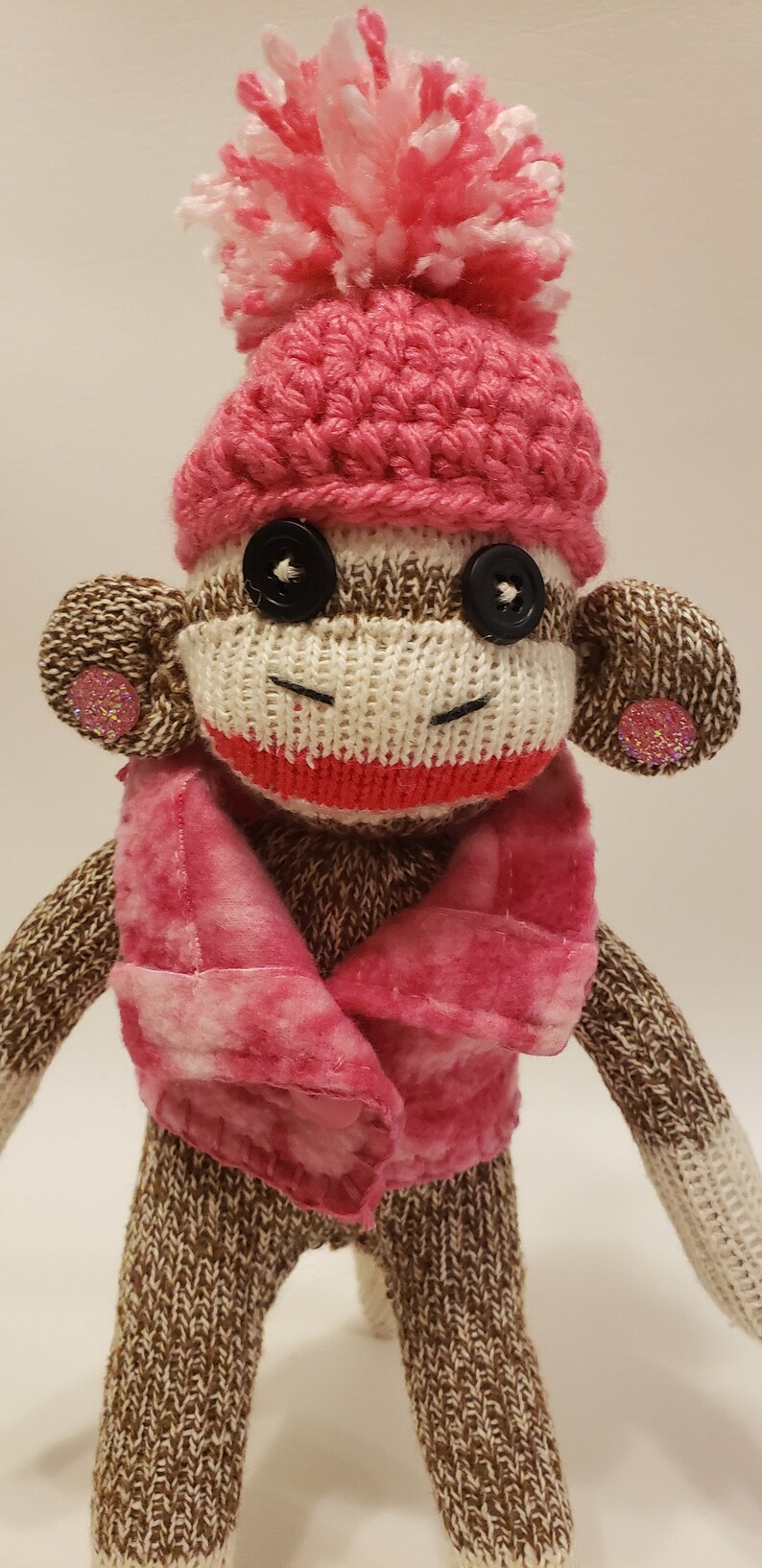 May include: A brown and white sock monkey wearing a pink crocheted hat with a white pom-pom and a pink and white plaid scarf.