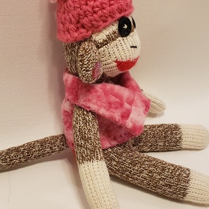 May include: A brown and white sock monkey wearing a pink crocheted hat with a white pom-pom and a pink fuzzy scarf.