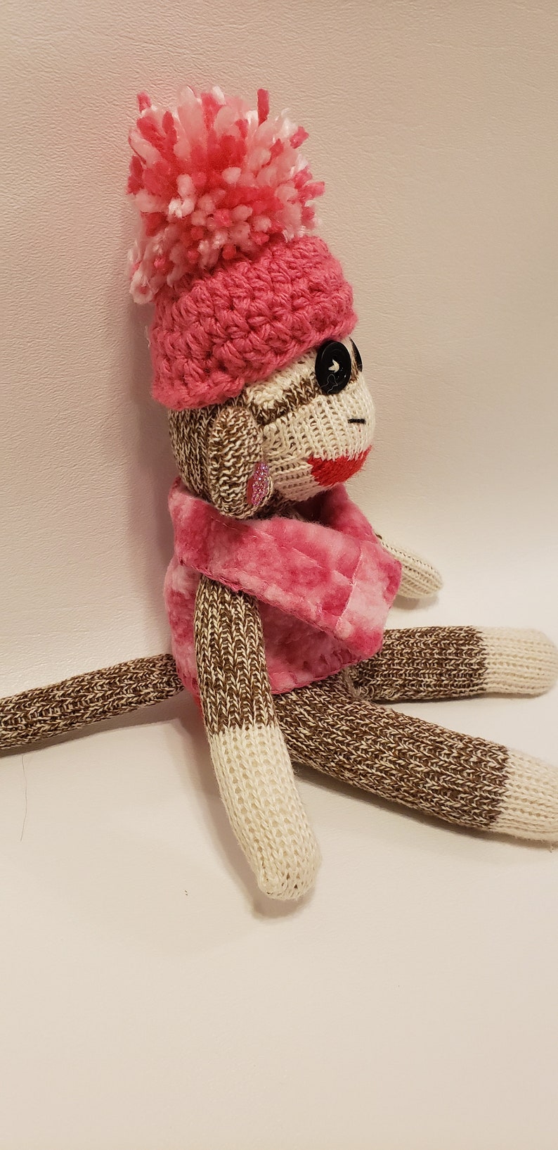 May include: A brown and white sock monkey wearing a pink crocheted hat with a white pom-pom and a pink fuzzy scarf.