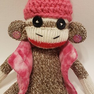 May include: A brown and white knitted sock monkey wearing a pink crocheted hat with a white pom-pom and a pink and white plaid jacket. The monkey has black button eyes and pink glitter buttons on its ears.