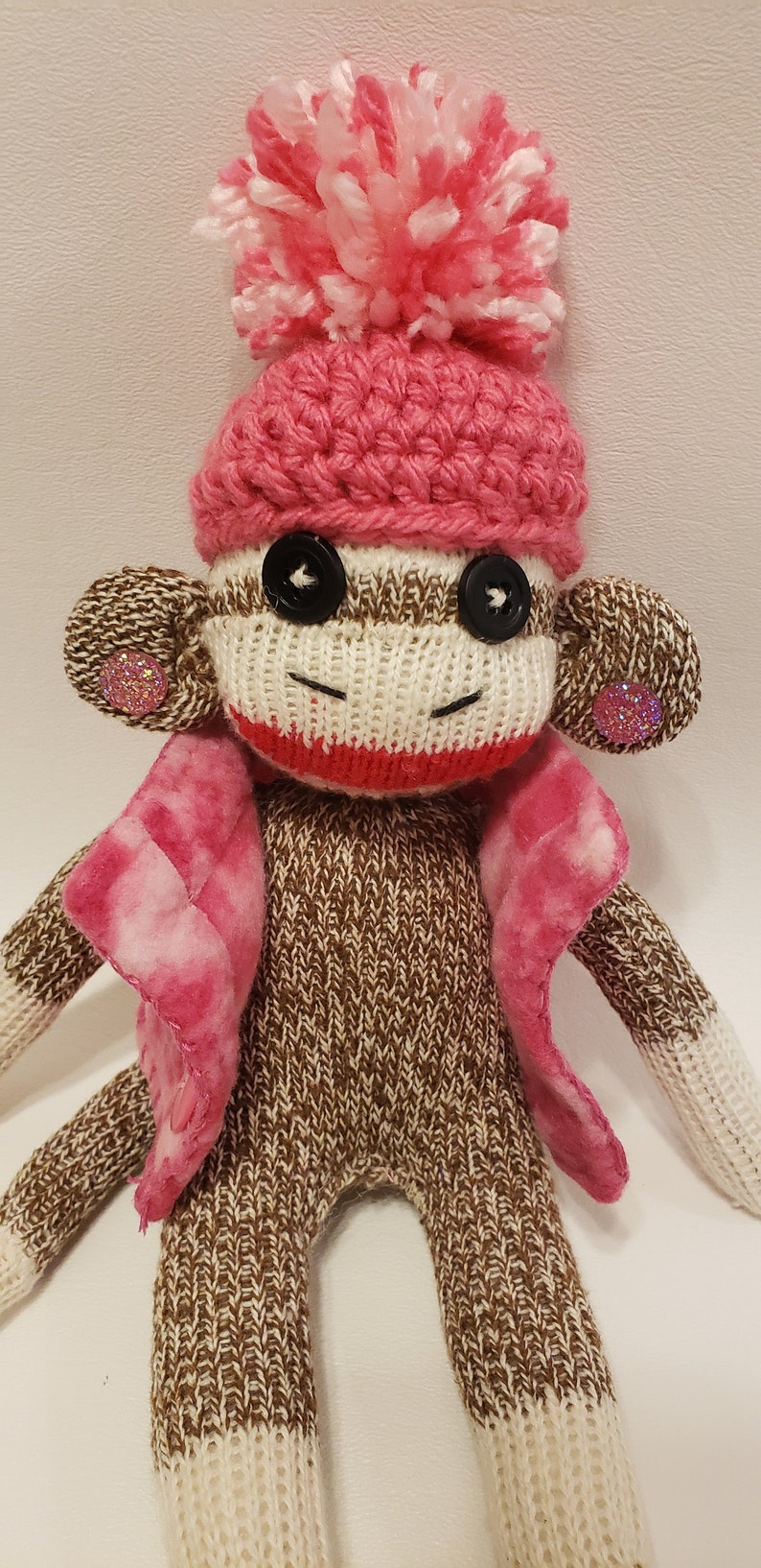 May include: A brown and white knitted sock monkey wearing a pink crocheted hat with a white pom-pom and a pink and white plaid jacket. The monkey has black button eyes and pink glitter buttons on its ears.