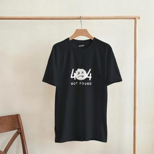 May include: Black t-shirt with a white graphic featuring the number 404, crossed-out eyes, and a sad mouth, with the words "NOT FOUND" below. The shirt is hanging on a wooden hanger.
