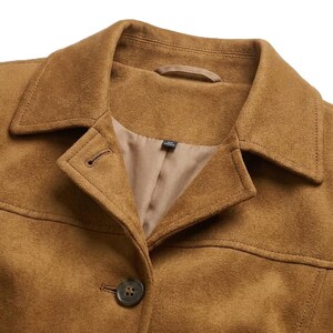 May include: A tan suede-textured jacket featuring a classic pointed collar, a single visible dark button closure, and a smooth interior lining. The garment includes a horizontal seam across the chest and a small fabric loop at the inner neckline.