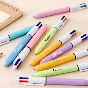 Peut inclure: Plusieurs stylos multicolores aux couleurs pastel, dont rose, bleu, jaune et vert, avec des détails blancs. Certains stylos sont personnalisés avec des noms tels que "Winnie", "Keith", "Chris", "Devin" et "Lucie".