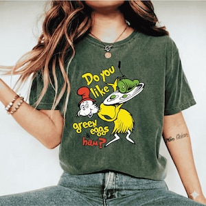 Puede incluir: Camiseta verde con una ilustración de dibujos animados de un personaje sosteniendo un plato con huevos verdes y jamón. El texto "Do you like green eggs & ham?" está impreso en amarillo y rojo.