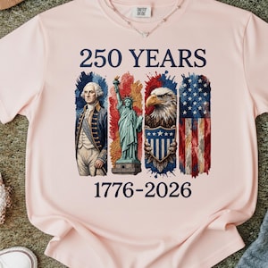 以下が含まれることがあります： 「250 YEARS」の文字と、ジョージ・ワシントン、自由の女神、ワシ、アメリカ国旗のイラストが描かれた、薄いピンク色のTシャツ。「1776-2026」の年号がイラストの下に印刷されています。