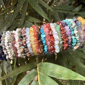 Può includere: Una collezione di braccialetti con schegge di pietre preziose in vari colori, tra cui blu, rosa, verde e arancione. I braccialetti sono esposti su una superficie cilindrica beige su uno sfondo di fogliame verde.