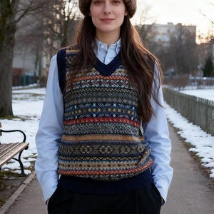 Puede incluir: Una persona con un gorro de piel marrón, un chaleco de punto estampado con ribete azul marino y una camisa celeste abotonada. El chaleco presenta un diseño geométrico en tonos marrón, azul y rojo. El fondo muestra un parque.