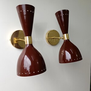 Puede incluir: Par de apliques de pared modernos de mediados de siglo. Los apliques tienen una pantalla en forma de cono rojo oscuro con pequeños puntos blancos cerca de la parte superior. La base y los herrajes de conexión son dorados. Los apliques están montados en una pared blanca.