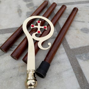 Könnte beinhalten: Ein hölzerner Gehstock mit einem goldfarbenen Griff und einem Kreuzdesign mit roten Akzenten. Der Stock hat eine dunkelbraune Oberfläche und eine schwarze Gummispitze. Der Griff ist verziert und detailliert.