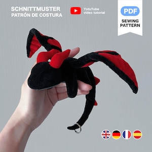 Peut inclure: Un jouet en peluche en forme de dragon noir et rouge avec de grandes ailes et une queue incurv&eacute;e. Le dragon a un anneau argent&eacute; attach&eacute; &agrave; sa queue. Le texte de l'image indique "SCHNITTMUSTER PATR&Oacute;N DE COSTURA" et "PDF SEWING PATTERN".