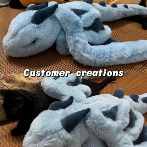 Peut inclure: Une peluche en forme de dragon bleu clair avec des d&eacute;tails bleu fonc&eacute;, comme les cornes et les motifs. Le jouet est doux et duveteux, avec un long corps et une queue. Le texte "Customer creations" est visible.