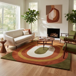 May include: A living room scene featuring a beige sofa, two green armchairs, and a round wooden coffee table. A large rug with concentric oval patterns in green, orange, yellow, and white dominates the floor. A painting hangs above the fireplace.