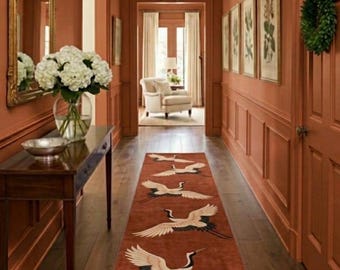 May include: A hallway with terracotta walls and dark wood flooring. A runner rug featuring crane designs in cream, black, and red runs down the centre. A dark wood console table with a mirror and a vase of white flowers is on the left.