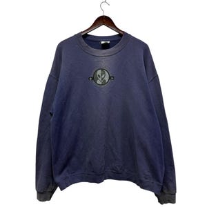 May include: A navy blue sweatshirt with a crew neck and long sleeves. The front features a circular graphic with an alien head design. The sweatshirt is hanging on a wooden hanger against a white background.