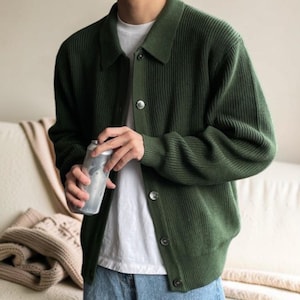 Puede incluir: Un cárdigan verde oscuro con botones y textura acanalada. El cárdigan tiene cuello y se lleva sobre una camiseta blanca. La persona sostiene una lata plateada. El cárdigan tiene botones plateados.