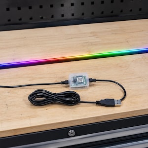 Peut inclure: Une barre lumineuse LED multicolore avec un câble noir, un petit boîtier de commande transparent et un connecteur USB. La bande lumineuse passe du bleu au rouge au vert. Les éléments sont sur une surface en bois.