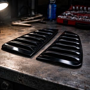 Puede incluir: Dos cubiertas de ventilación automotriz negras con lamas horizontales, sobre una superficie metálica texturizada. Las cubiertas son brillantes y reflejan el entorno. La imagen está tomada en un taller, con herramientas y una lámpara visibles. Miden unos 30 cm.