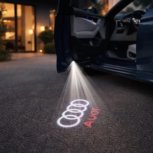 May include: A dark blue car with the door open, illuminating the ground with a bright light. The light projects the Audi logo and the brand name in red onto the asphalt driveway.