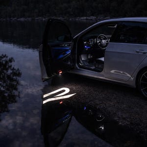 Peut inclure: Une voiture blanche avec la portière ouverte, projetant un logo sur le sol. Les lumières intérieures sont allumées et la voiture est garée près de l'eau. La scène est sombre, les lumières de la voiture étant la principale source d'éclairage.