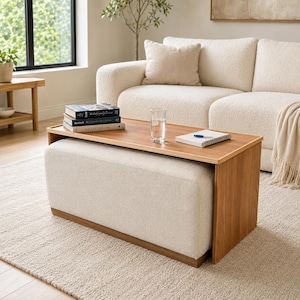 May include: A rectangular ottoman featuring a cream-colored textured fabric base and a smooth wooden top surface. The wooden top extends over the sides, creating a functional table surface holding a stack of three books, a glass of water, and a notebook.