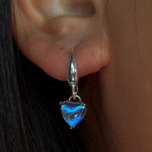 May include: A silver earring with a heart-shaped, iridescent blue charm. The charm displays a gradient of blue hues. The earring is attached to the ear with a silver hook. The background is a person's hair and skin.