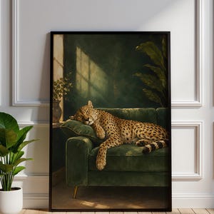 May include: A framed wall art print featuring a leopard with golden fur and black spots sleeping on a dark green velvet sofa. The scene includes a potted plant on a side table and large green leaves in the background, all set in a room with sunlight.