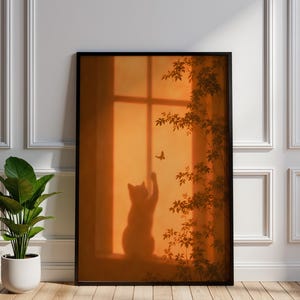 Può includere: Una stampa artistica rettangolare in una cornice nera che mostra la silhouette di un gatto seduto sul davanzale di una finestra che cerca di afferrare una farfalla. La scena è ambientata su uno sfondo di tramonto arancione con ombre di rami.
