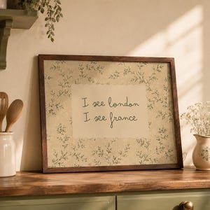May include: A rectangular wall art print in a dark brown wooden frame featuring a beige background with a delicate green botanical vine pattern. The center displays the black text "I see london I see france" in a simple, handwritten-style font.