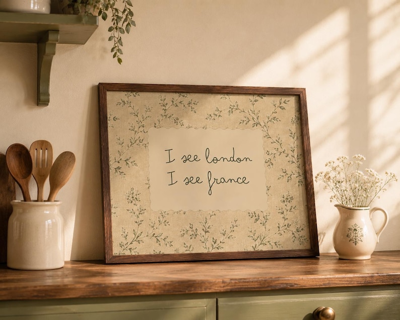 May include: A rectangular wall art print in a dark brown wooden frame featuring a beige background with a delicate green botanical vine pattern. The center displays the black text "I see london I see france" in a simple, handwritten-style font.
