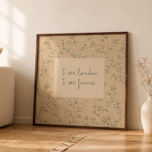 May include: A square framed wall art piece featuring a beige background with a delicate dark green botanical vine pattern. In the center, a cream-colored rectangular inset displays the black handwritten text: I see london I see france.