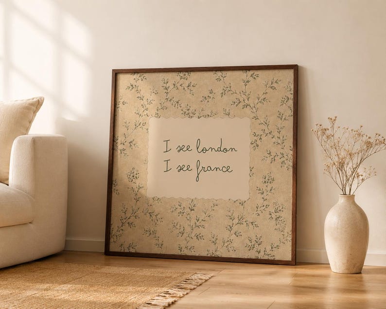 May include: A square framed wall art piece featuring a beige background with a delicate dark green botanical vine pattern. In the center, a cream-colored rectangular inset displays the black handwritten text: I see london I see france.