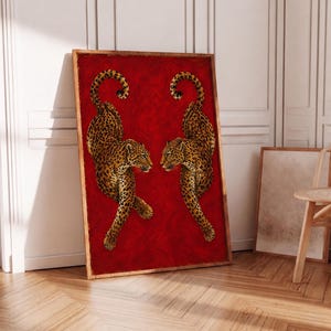 Puede incluir: Obra de arte enmarcada con dos leopardos enfrentados sobre un fondo rojo. Los leopardos tienen un patrón de manchas en tonos marrones y negros. La obra está en un marco de madera y se apoya contra una pared blanca.