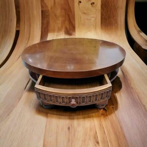 Pode incluir: Uma mesa redonda de madeira com uma gaveta. A mesa é de cor castanho médio com um acabamento brilhante. A gaveta está aberta, revelando o interior. A mesa está sobre um fundo de madeira.