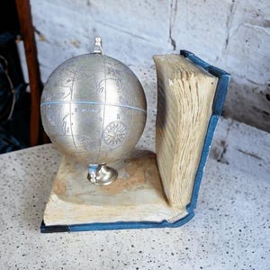 May include: A silver-colored globe sits atop a book-shaped stand with a map design. The bookends are blue with aged-looking pages. The globe features engraved continents and a compass rose. A decorative item for a desk or shelf.