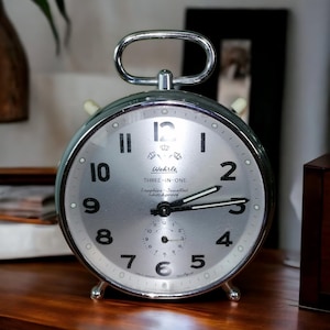May include: A silver-toned Wehrle Three-in-One alarm clock with a round face and a handle. The clock face has black numerals and hands, with the words "Wehrle Three-in-One Sapphire Jewelled Shockproof" printed on the face. The clock is on a wooden surface.