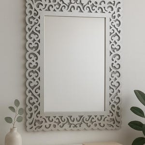 以下が含まれることがあります： 装飾的なスクロールワークフレームが付いた長方形のシルバーミラー。白い壁に掛けられており、白い表面の上にあります。小さな花瓶と本が表面に置かれています。
