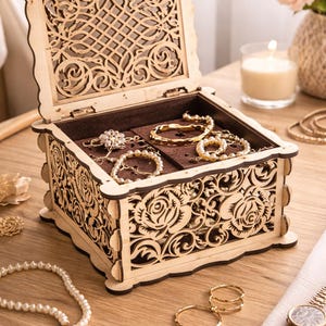 May include: Open wooden jewelry box with intricate laser-cut floral and geometric designs. The box contains various gold bracelets, rings, and a pearl necklace. A gold watch and a lit candle are also visible.