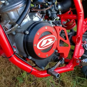 Op de afbeelding: Close-up van een motorfietsmotor met een rood frame en accenten. De motorbehuizing is zwart met een rood en wit logo. De componenten van de motorfiets omvatten een ketting, een voetsteun en diverse mechanische onderdelen.