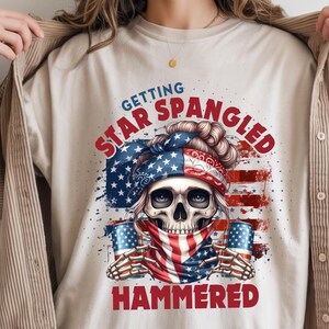 Puede incluir: Una camiseta beige con un gráfico de una calavera que lleva un pañuelo con el patrón de la bandera estadounidense, una braga de cuello a juego y sostiene dos latas de bebida. El texto "GETTING STAR SPANGLED HAMMERED" aparece en rojo y azul.