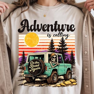 Könnte beinhalten: Ein beiges T-Shirt mit einer Grafik eines türkisfarbenen Geländewagens mit einem Skelett am Steuer. Der Text lautet "Adventure is calling" und "On the Road Again" auf dem Reserverad. Der Hintergrund zeigt eine gelbe Sonne, Tannen und Berge.