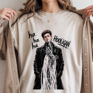 Op de afbeelding: Beige t-shirt met een afbeelding van een persoon in een zwarte jas en een skelet hand die een microfoon vasthoudt. Het woord "Hooligan" is in het zwart geschreven, met "ha ha ha" boven de afbeelding.