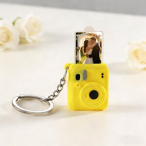 May include: A yellow camera-shaped keychain with a silver chain and ring. The camera has a black lens and a small photo of a couple embracing. The photo is displayed in the top slot of the camera.