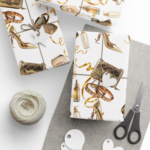Puede incluir: Papel de regalo blanco con un patrón repetido de tacones dorados, bolsos, frascos de perfume y joyas. Los regalos envueltos están atados con hilo y adornados con etiquetas en forma de estrella. Cintas doradas se enrollan alrededor de los regalos.